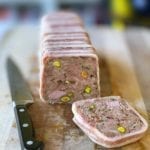 A terrine of country pate with one slice cut and resting on a wooden cutting board with a knife beside it