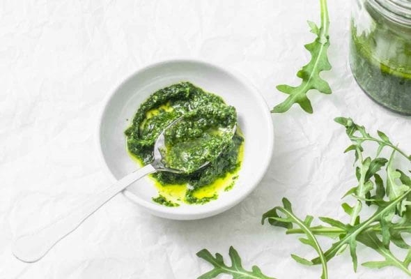 A white bowl partially filled with arugula pesto and a spoon resting in the pesto. A few arugula leaves scattered beside the bowl.
