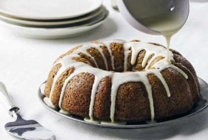 An old-fashioned zucchini cake on a silver plate with lemon glaze being drizzled over it.