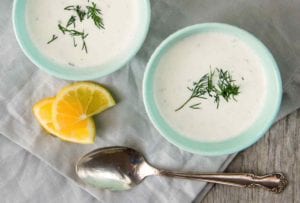 Two blue bowls of cold cucumber soup with yogurt and dill on a linen cloth with a spoon and lemon beside them.