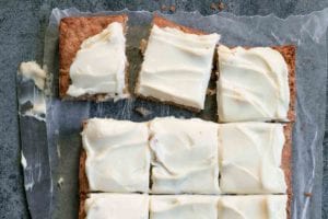 A frosted apple cake on a sheet of wax paper that has been cut into 12 squares with a knife resting beside.