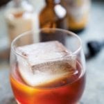 A high ball glass partially filled with an amber Sazerac cocktail without absinthe and an oversized ice cube.
