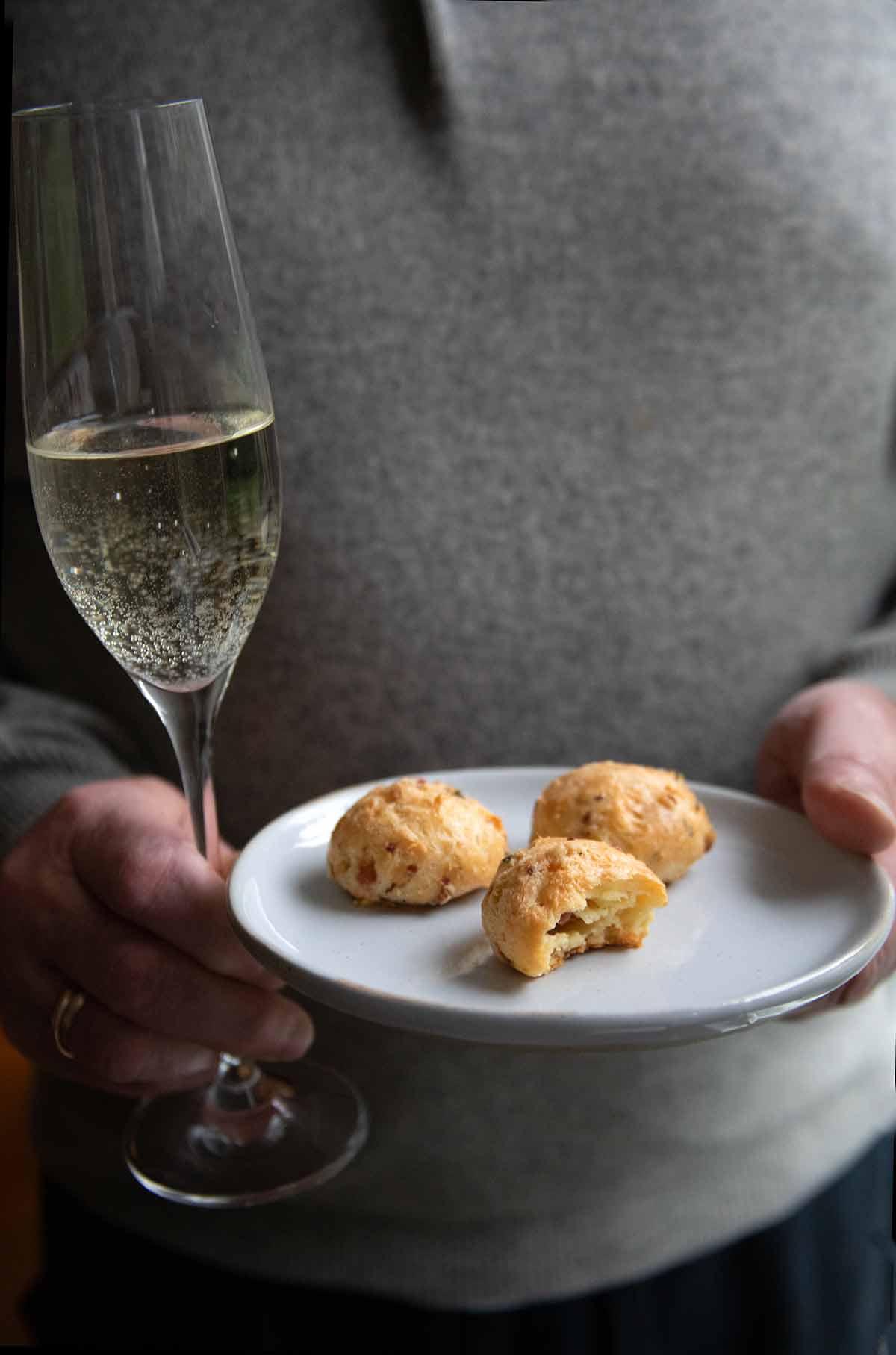 A man holding a glass of Prosecco and a plate with three gougeres.