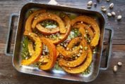 A metal roasting pan filled with roast butternut squash, feta, and pistachios on a wooden table with pistachios scattered around it.