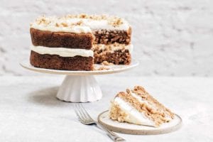 A whole two layer spiced carrot cake on a white cake stand with one slice cut from it on a plate with a fork beside it.