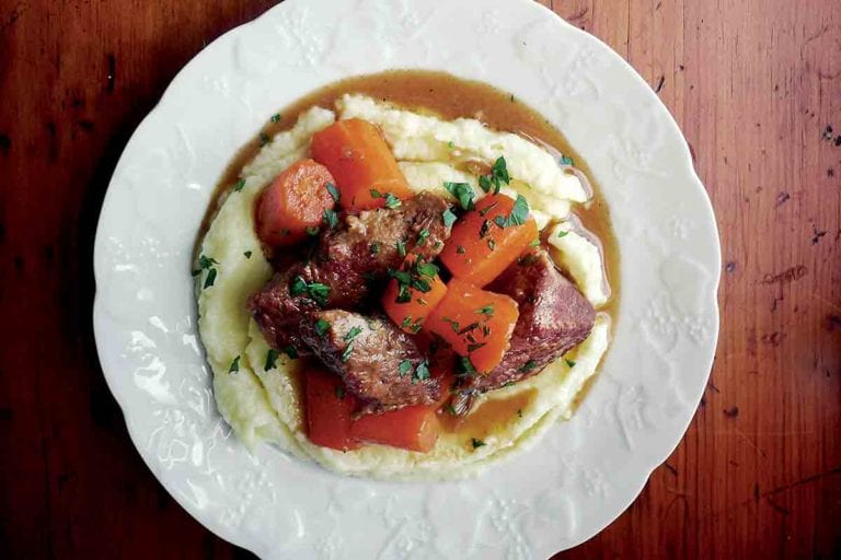 Two white plates of braised beef with carrots on top of mashed potatoes.
