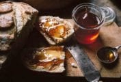 Two loaves of artisan bread and a jar of Campari citrus marmalade, with a slice of bread smeared with the marmalade on a wooden cutting board.