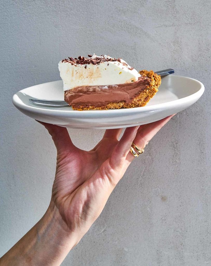 A person holding a white plate with a slice of dark chocolate pudding pie and a fork.
