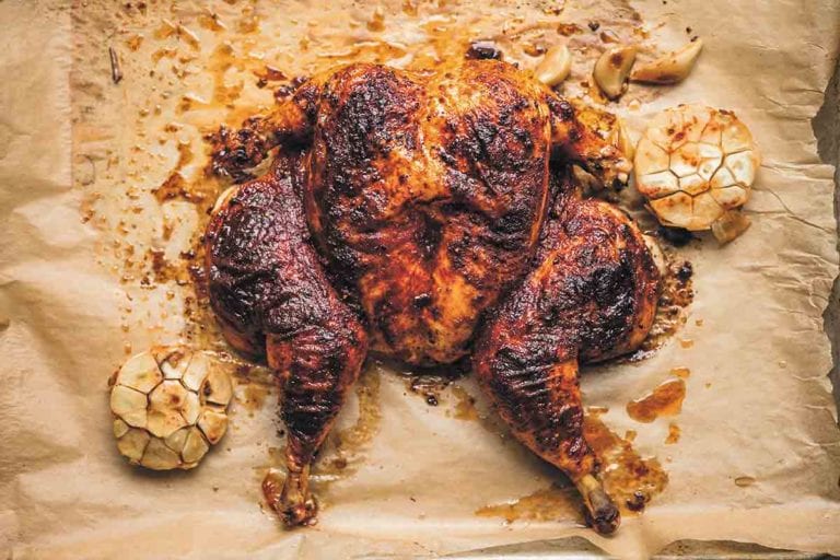 A flat roasted chicken with smoked paprika on a sheet of parchment paper with a halved head of garlic on either side.