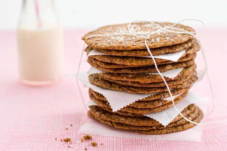 A stack of thin and crisp chocolate chip cookies with pieces of parchment in between, tied up with string and a glass bottle of milk in the background.