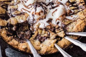 A chocolate chunk skillet cookie topped with ice cream and chocolate sauce in a cast-iron skillet with four spoons resting in the cookie.