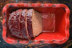 Half a cooked meatloaf in a red loaf tin with two slices cut from it.