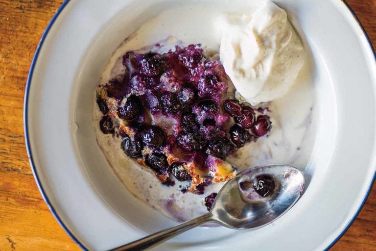 A white bowl with a serving of blueberry clafouti, a scoop of vanilla ice cream, and a spoon resting in the bowl.