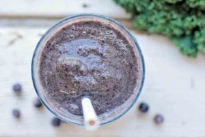 An overhead view of a glass of blueberry kale smoothie with a straw and some blueberries and kale scattered around the glass.