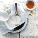 Homemade Greek yogurt being strained through cheesecloth into a white bowl with a spoon and a small bowl of honey beside it.