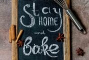 A small wood-rimmed chalkboard with "Stay Home and Bake" written on it, with a whisk, eggshell, and cinnamon and star anise scattered beside it.