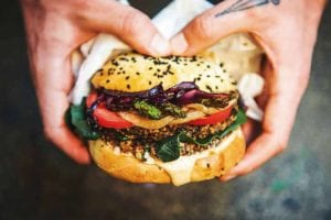 A person holding a black bean burger filled with spinach, tomato, onion, and asparagus.