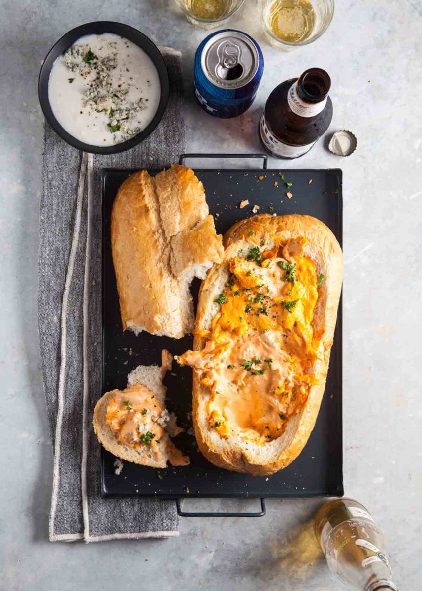 A loaf of Italian bread filled with buffalo chicken dip, with a dish of blue cheese dip and a few beers on the side.