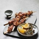 Four grilled pork skewers on a white plate with a bowl of lemon wedges and dried oregano beside the skewers.