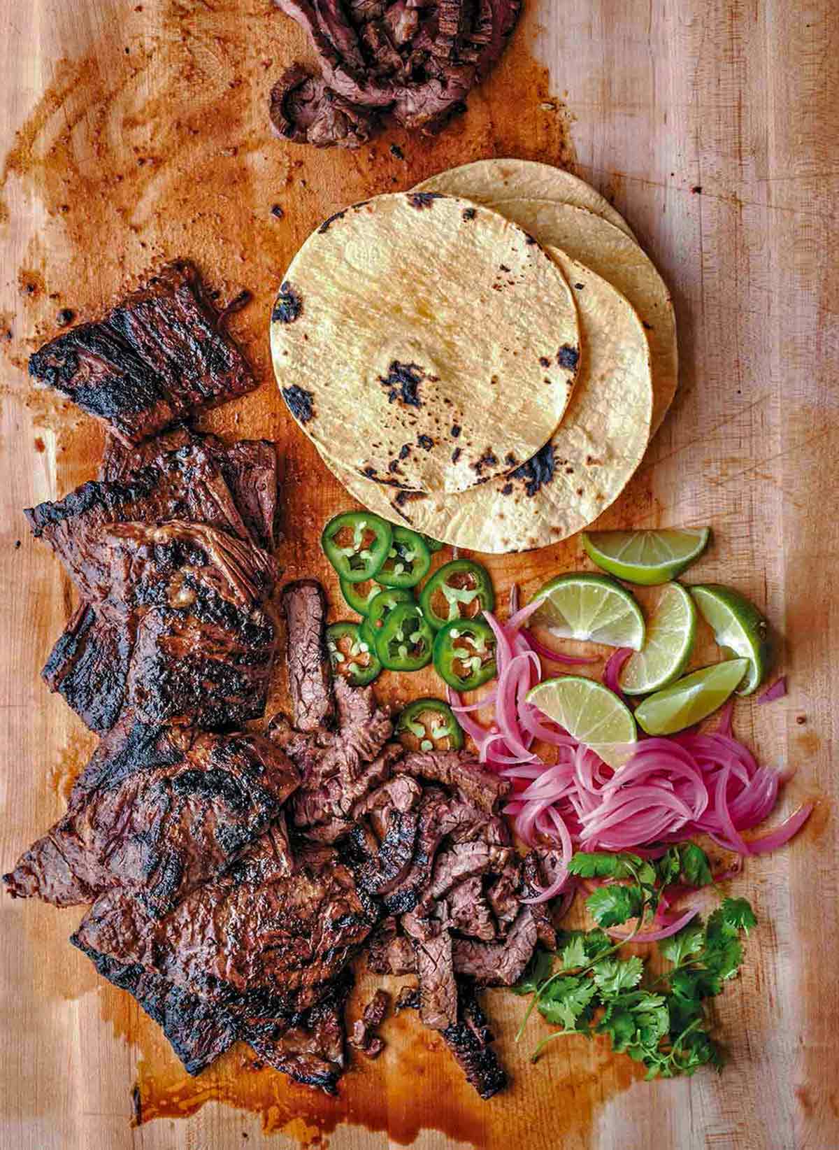 Cutting board with grilled skirt steak tacos, sliced jalepenos, tortillas, lime wedges, pickled onions, cilantro