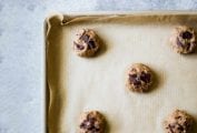 A parchment-lined rimmed baking sheet with balls of uncooked chocolate chip cookie dough in answer to the question 'why are my chocolate chip cookies flat?'.