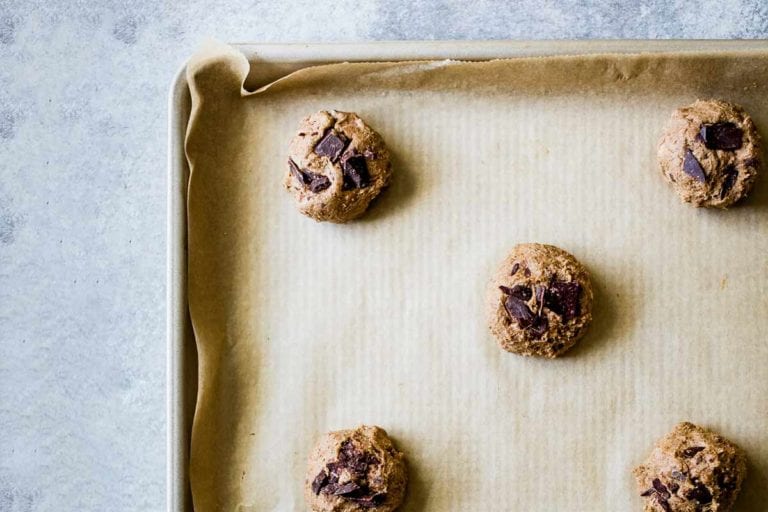 A parchment-lined rimmed baking sheet with balls of uncooked chocolate chip cookie dough in answer to the question 'why are my chocolate chip cookies flat?'.