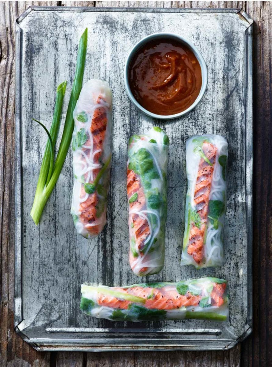 Four salmon summer rolls on a metal sheet with a piece of scallion and a bowl of dipping sauce.