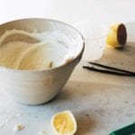 A bowl of sweet whipped ricotta cream, a halved lemon, two vanilla beans, a knife, and a small glass on a marble surface.