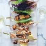Six chicken caesar salad skewers on a white plate with leaves of red and green romaine and two lemon wedges.