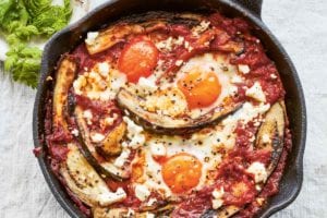 A cast-iron skillet filled with eggplant shakshuka made with poached eggs in a tomato and eggplant sauce, with feta scattered over the top.