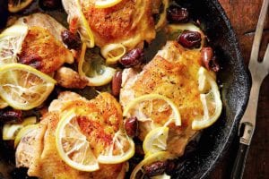 Four roast chicken thighs with lemon in a cast-iron skillet set on a wooden board with a meat fork resting beside it.