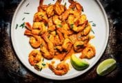 A white plate filled with masala shrimp, with lime wedges on the side, and gives sprinkled over, to garnish.