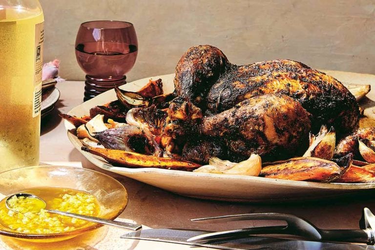 A serving platter topped with baharat roast chicken with sweet potatoes and a bowl of preserved lemon oil on the side along with a bottle of wine and a wine goblet.