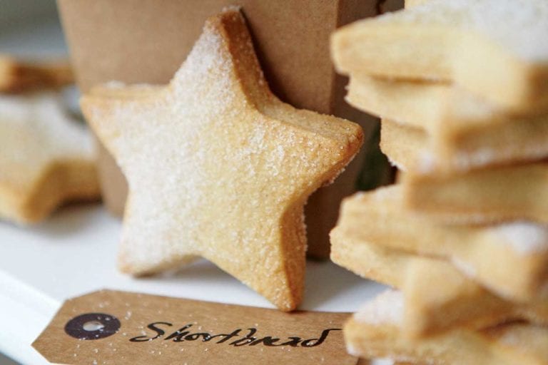 A stack of star-shaped cutout classic shortbread cookies, lightly dusted with sugar, and a brown takeout container and a gift tag