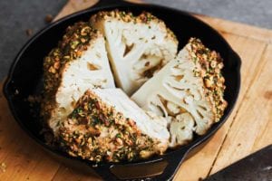 A whole roasted cauliflower with tahini and pistachios cut into four pieces in a cast-iron skillet.