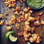 Florets of spicy roasted cauliflower on a baking sheet with squeezed lime wedges and a bowl of green sauce beside them.