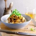 A white bowl filled with beef stroganoff and rice and topped with microgreens on a wooden board with a knife beside it.