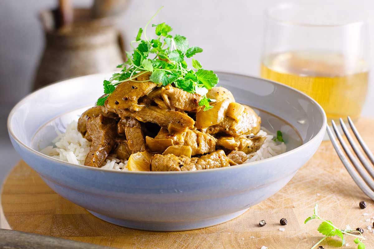 A white bowl filled with beef stroganoff and rice and topped with microgreens on a wooden board with a knife beside it.