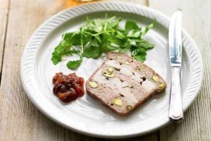 A white plate topped with a slice of chicken, pork, and pistachio terrine, a dollop of chutney, some arugula leaves and a knife.