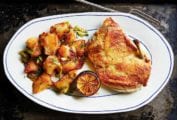 A piece of crisp roast chicken in a skillet on a plate with crispy potatoes and charred lemon.