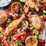 Greek-style sheet pan chicken, olives, zucchini, and lemon slices on a sheet pan with a bowl of tzatziki nearby.