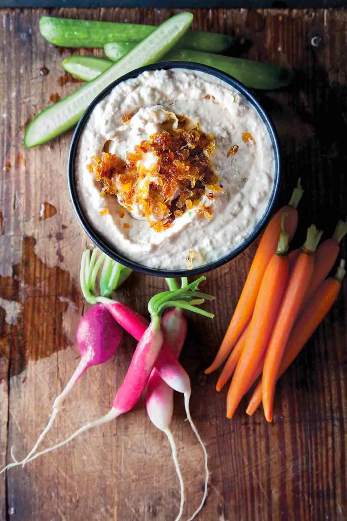 A bowl of onion dip surrounded by carrots, cucumbers, and radishes on a wooden board.