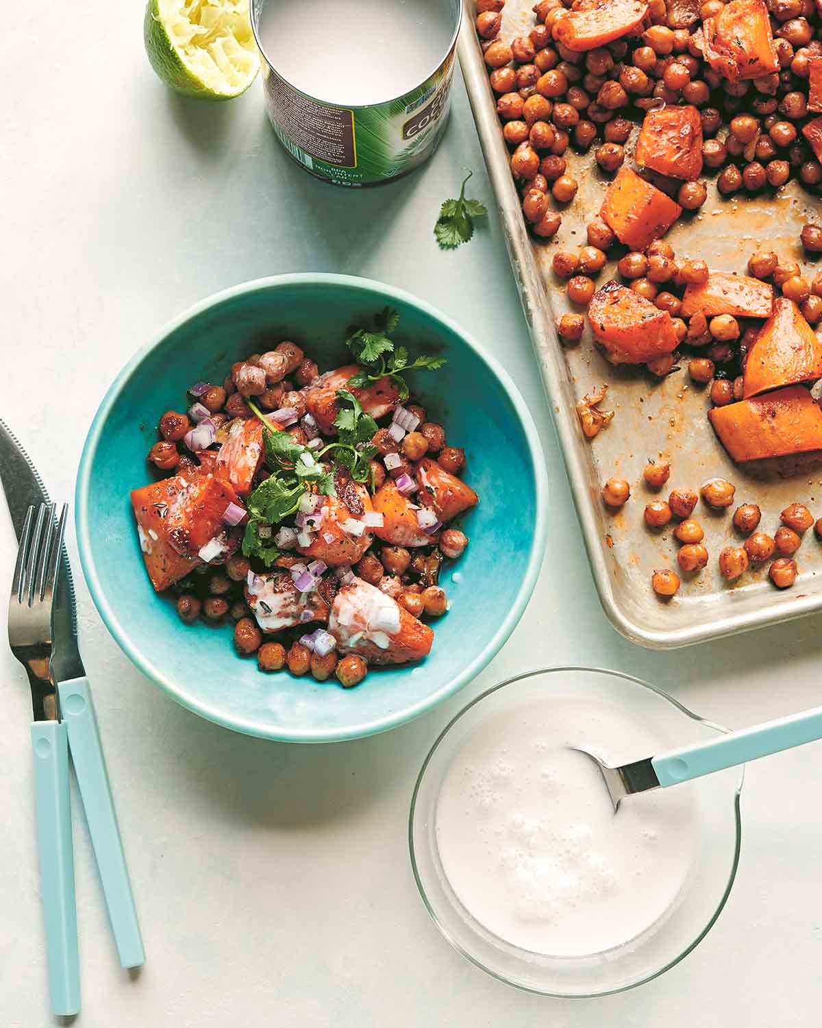 A bowl of roasted sweet potatoes and chickpeas, garnished with cilantro, next to a sheet pan and a bowl of coconut milk.