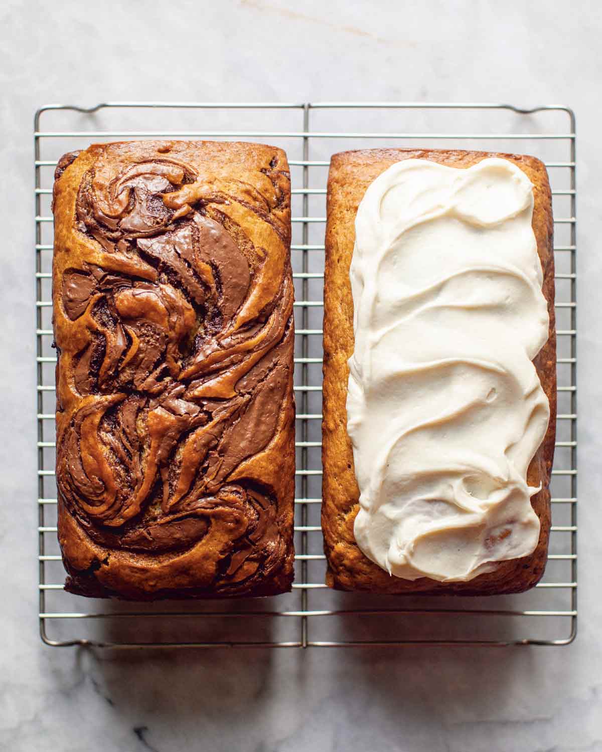 A loaf of banana bread with swirls of nutella next to a loaf of banana bread with cream cheese frosting on a wire rack.