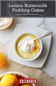 A lemon buttermilk pudding cake in a white bowl on a white plate, topped with whipped cream with a bowl of cream and basket of lemons on the side.