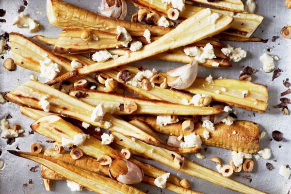 Halved roasted parsnips with hazelnuts and feta scattered on a baking sheet.