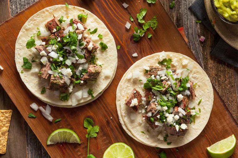Two steak tacos on a wooden board with lime wedges, onion, and cilantro scattered around.