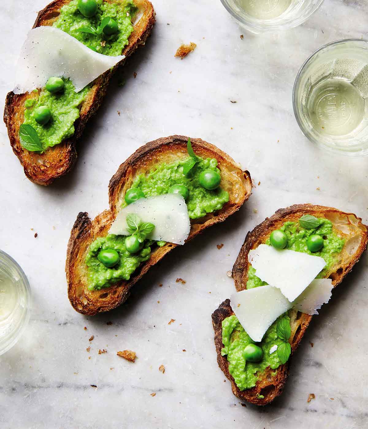 Three sweet pea crostini topped with pecorino shavings on a marble surface with glasses of white wine nearby.