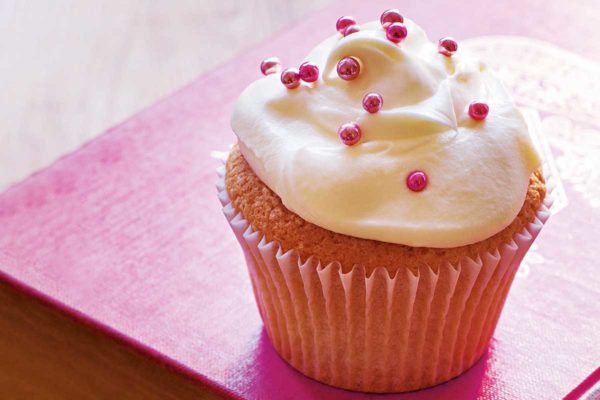A vanilla cupcake decorated with white frosting and pink sprinkles on a red book.