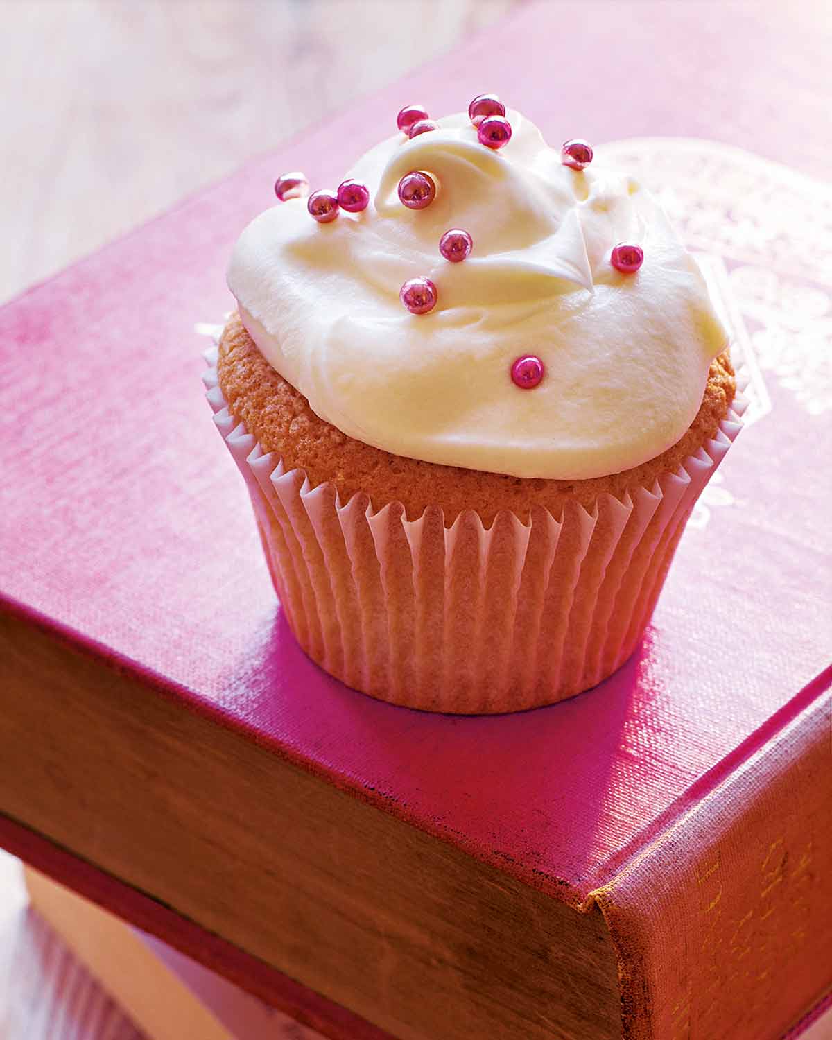 A vanilla cupcake decorated with white frosting and pink sprinkles on a red book.
